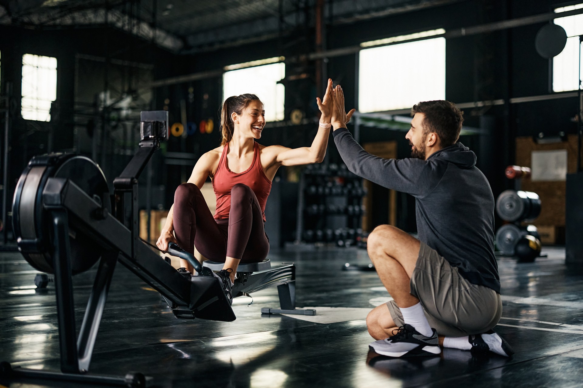 Happy athlete giving high-five to her personal trainer after exercising on rowing machine in a gym.